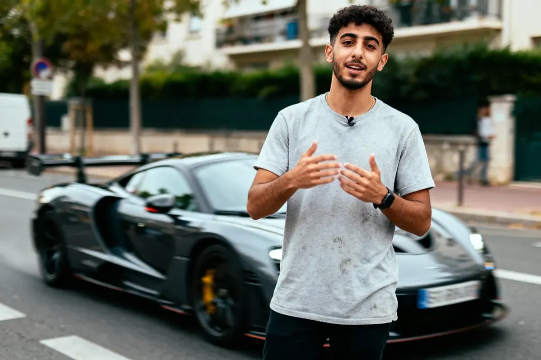 Un homme en t-shirt gris devant une voiture de sport noire garée dans la rue.