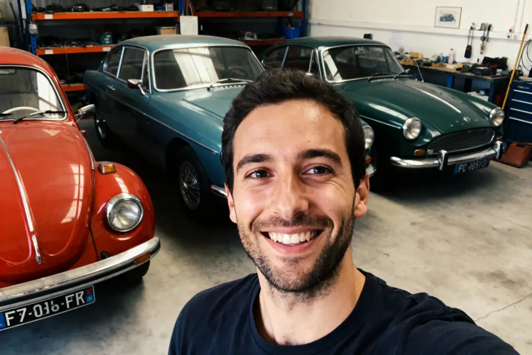 Homme souriant dans un garage avec trois voitures anciennes, dont une rouge et deux bleues.