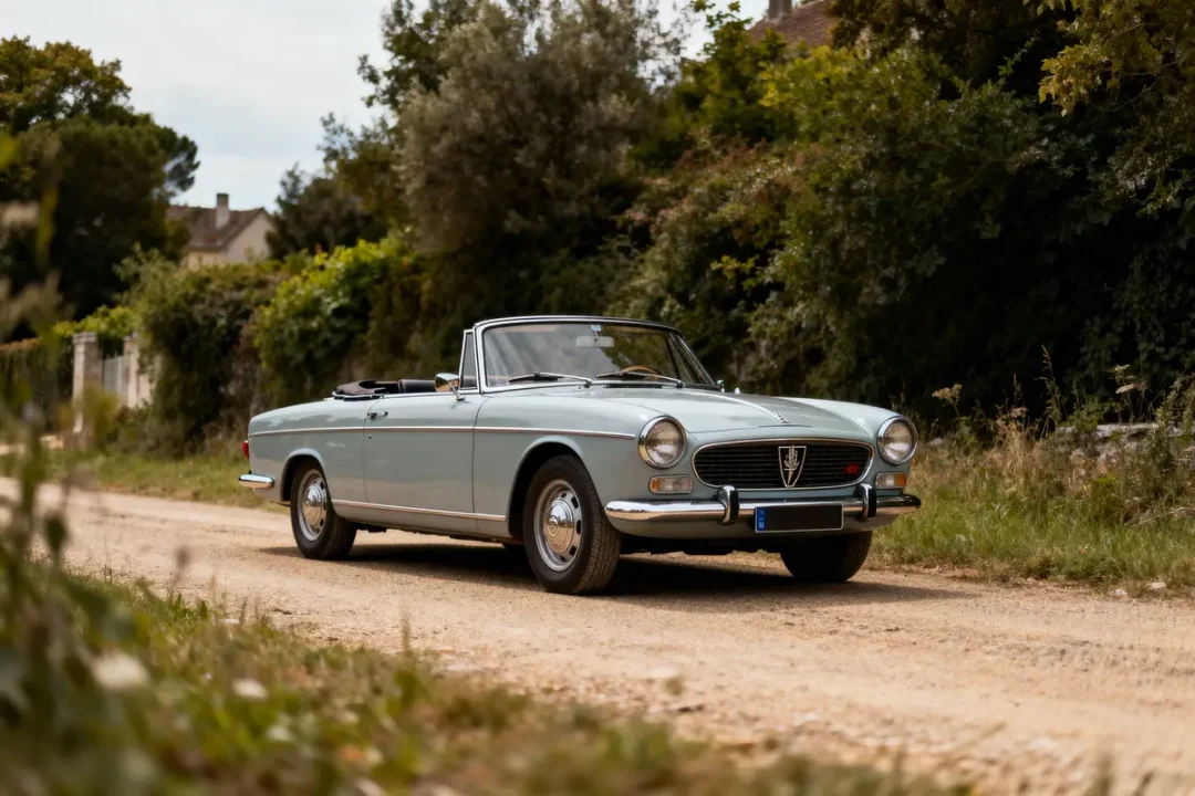 Voiture décapotable classique grise sur chemin de terre, entourée de végétation dense et ciel partiellement nuageux.