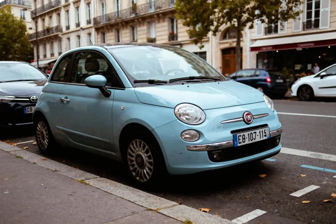 Petite voiture bleue Fiat stationnée dans une rue de la ville, bâtiments en arrière-plan, journée d'automne.