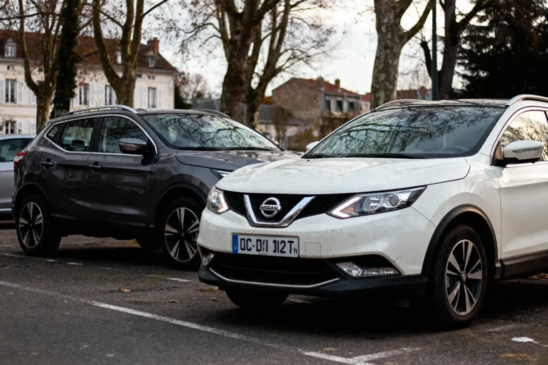 Deux voitures Nissan garées, l'une blanche et l'autre noire, sur un parking bordé d'arbres en hiver.