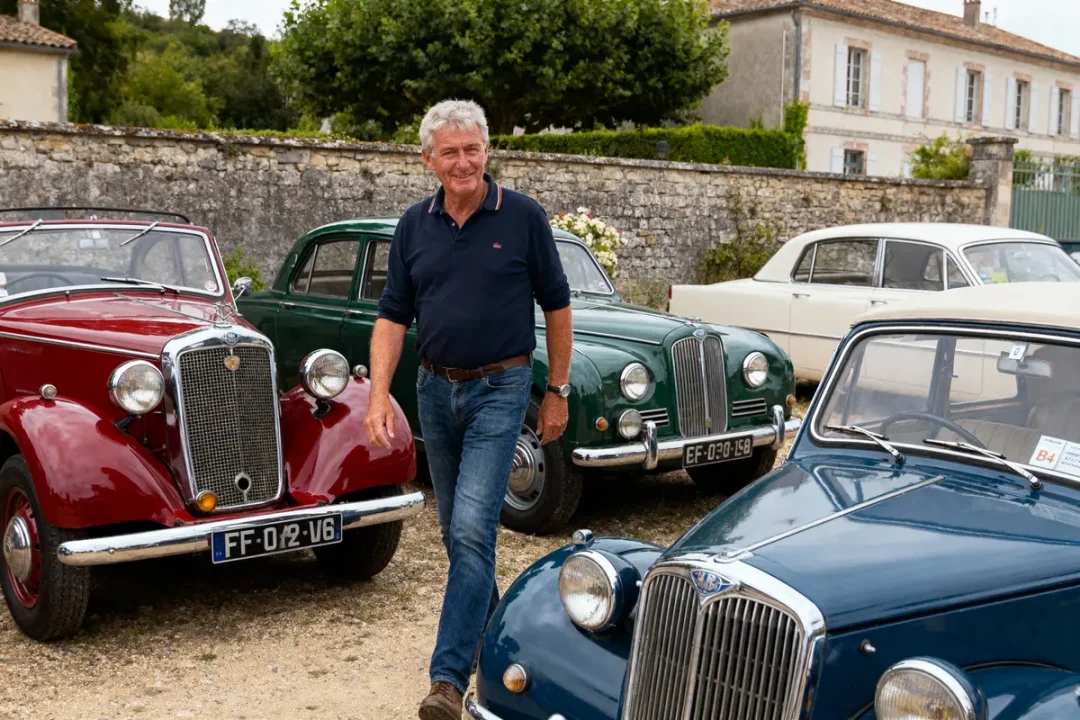 Un homme souriant marche devant des voitures anciennes colorées, en plein air, avec des bâtiments historiques en arrière-plan.