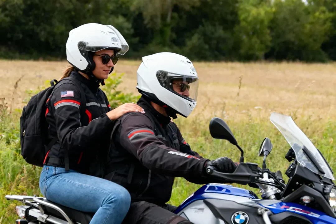 Deux personnes en moto avec casques blancs, roulant à travers un paysage champêtre verdoyant.