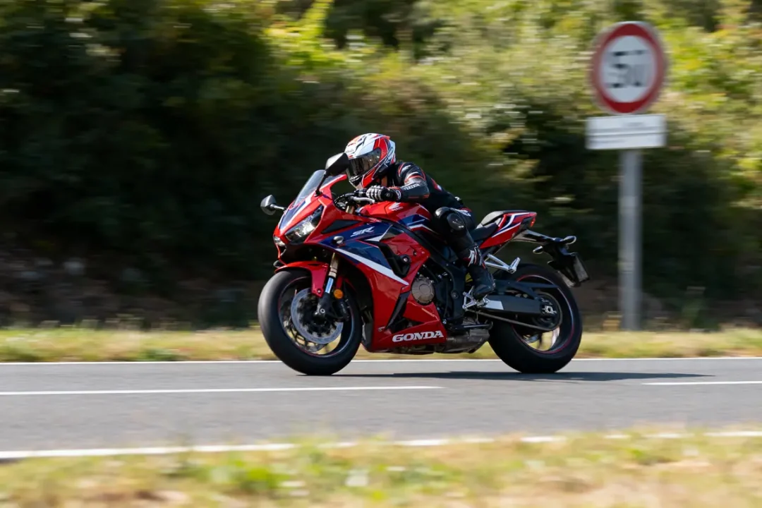 Motard en combinaison noire sur moto rouge, roulant vite sur route avec panneau de limitation de vitesse.