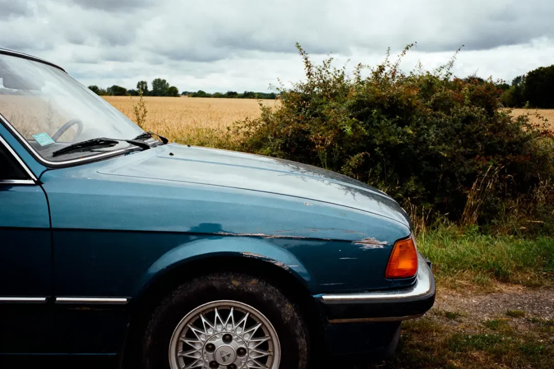Voiture bleue ancienne, champ de blé, buissons, ciel nuageux, campagne.