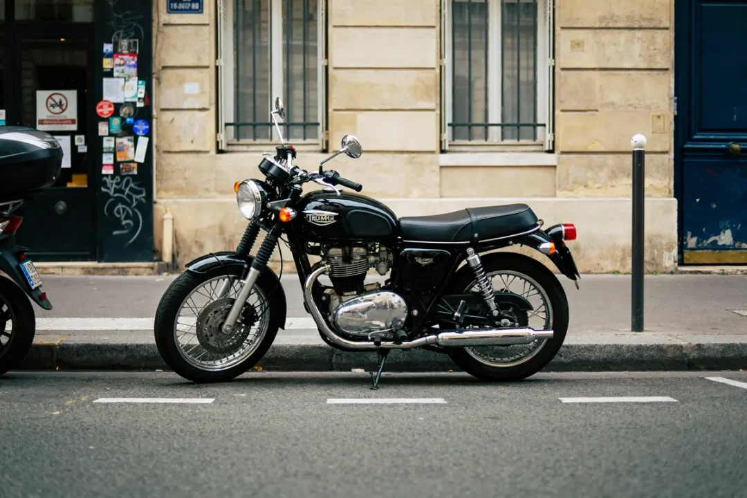 Une moto Triumph noire garée devant un bâtiment en pierre avec fenêtres grillagées, à côté d'un trottoir.