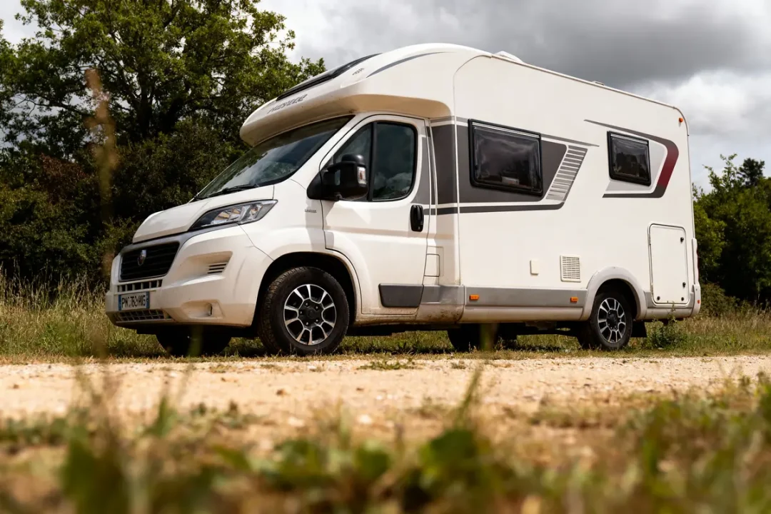 Camping-car blanc garé sur un chemin de terre, entouré de verdure, sous un ciel nuageux.