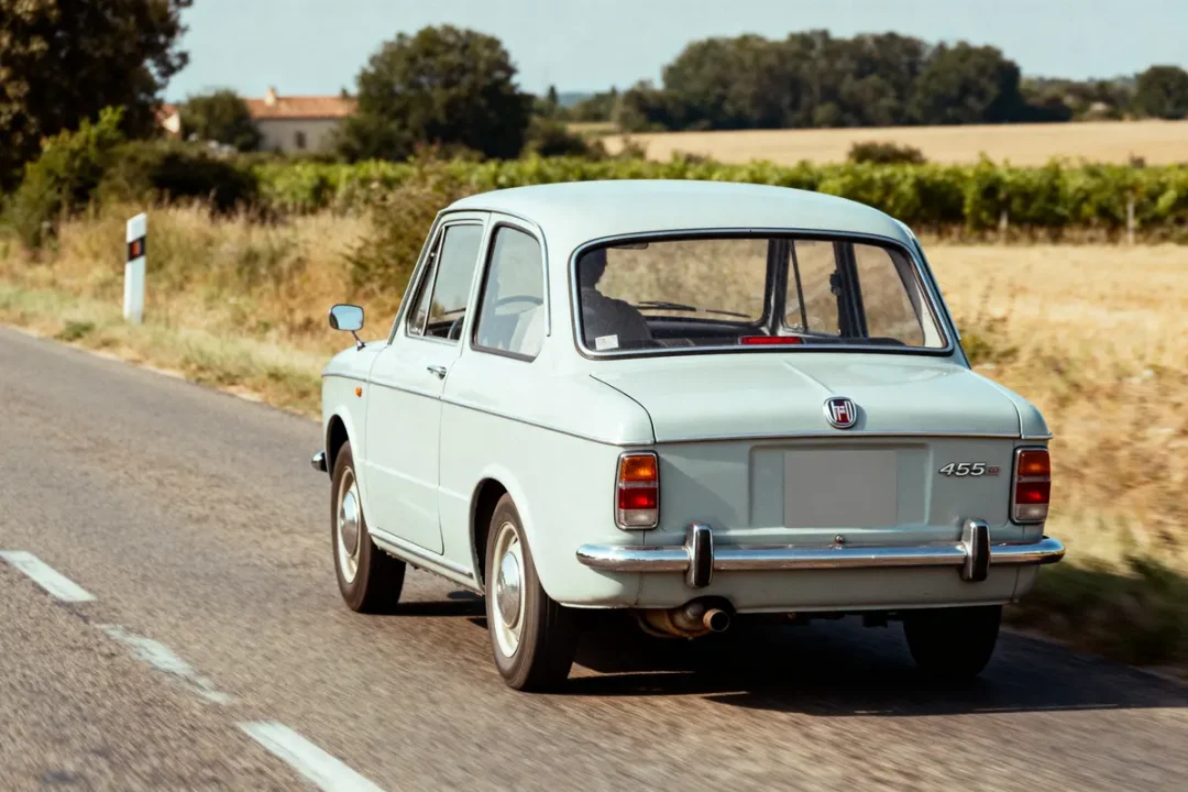 Voiture rétro bleue sur route de campagne, arbres et champs en arrière-plan, ciel dégagé.