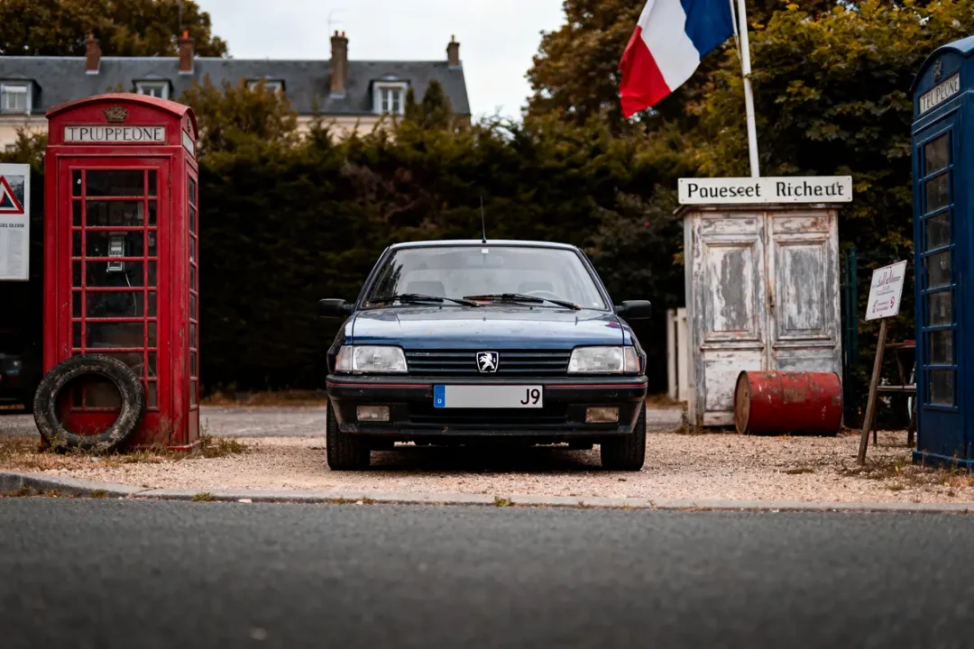 Voiture Peugeot bleue stationnée entre cabines rouges et bleues, drapeau français en arrière-plan.