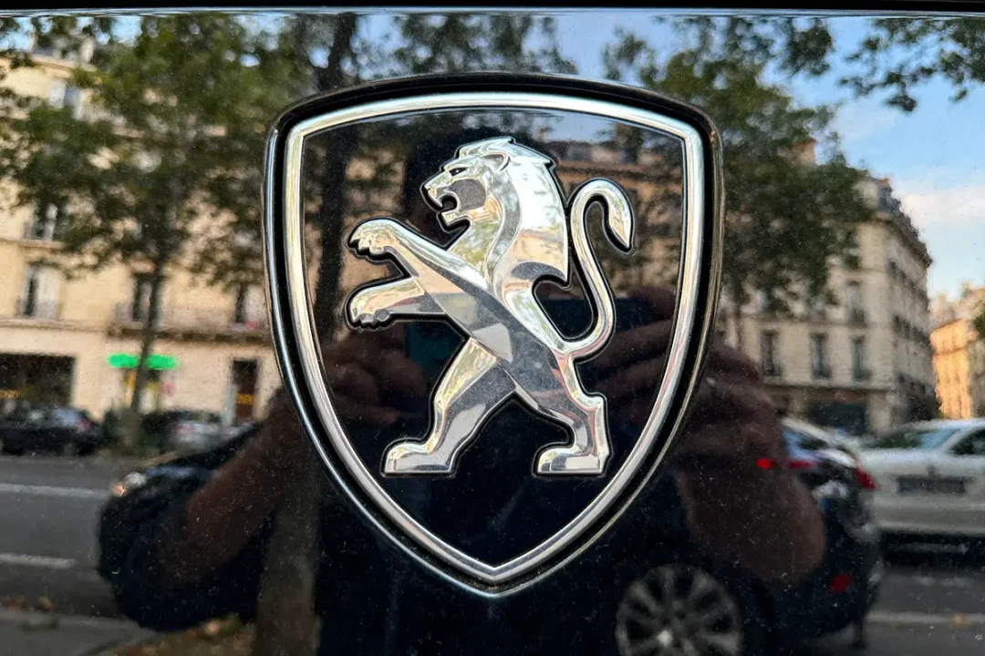 Logo argenté de lion Peugeot sur fond noir, reflet d'une rue urbaine en arrière-plan.