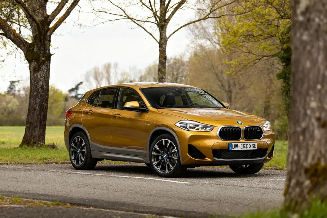 BMW X2 car dorée garée sur route entourée d'arbres en campagne verdoyante