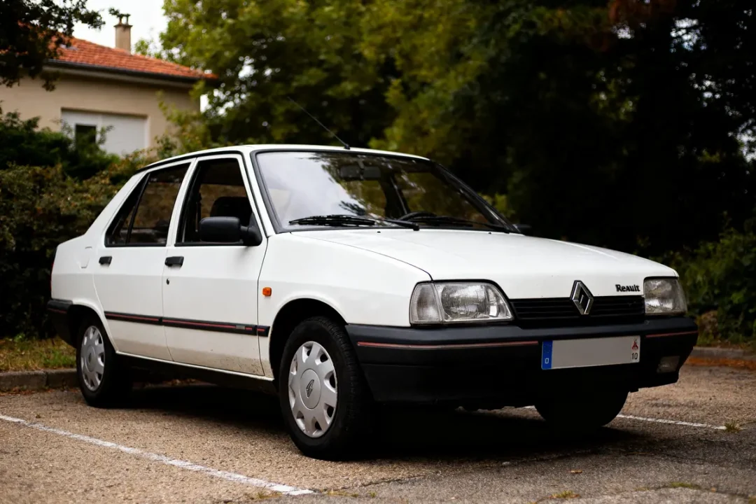 Voiture Renault 21 Sedan blanche garée sur un parking entouré de verdure