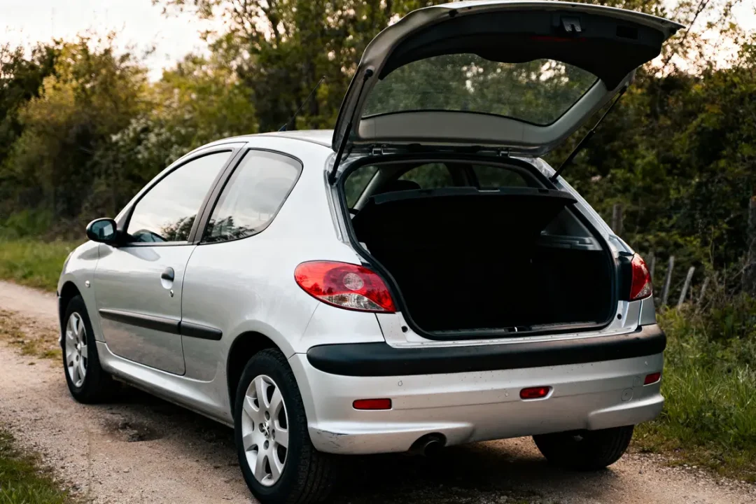 Hatchback Peugeot 206 argentée avec coffre ouvert sur chemin en terre.