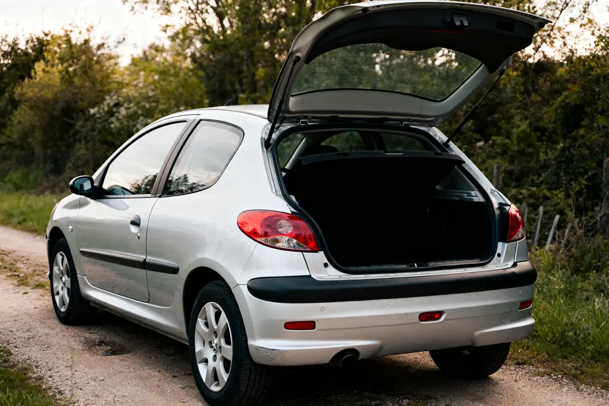 Hatchback Peugeot 206 argentée avec coffre ouvert sur chemin en terre.